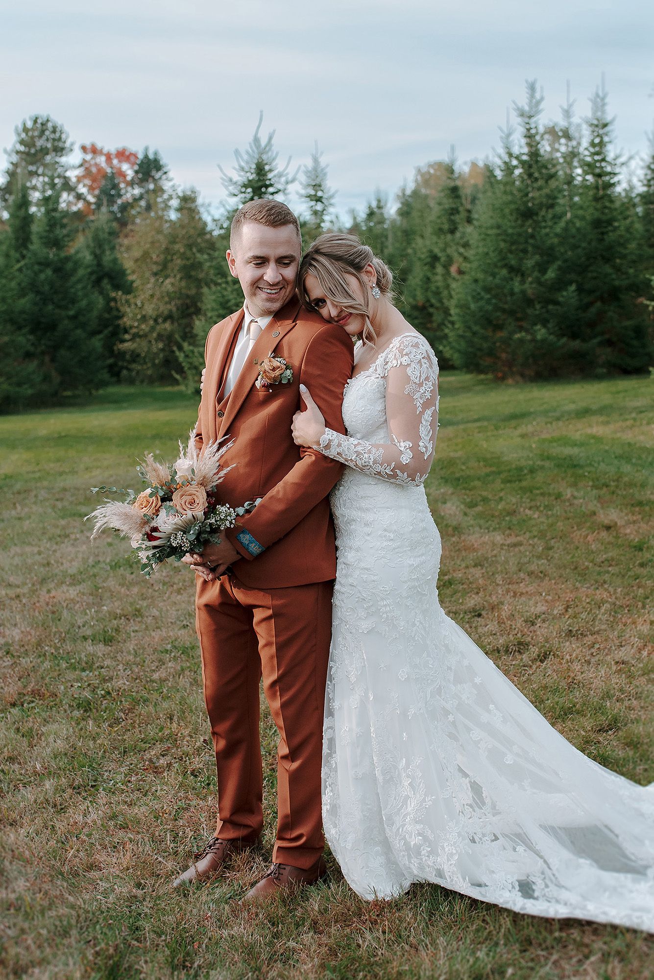 Wedding couple in rustic outdoor setting with bride in lace gown and groom in brown suit embracing on grassy hill.