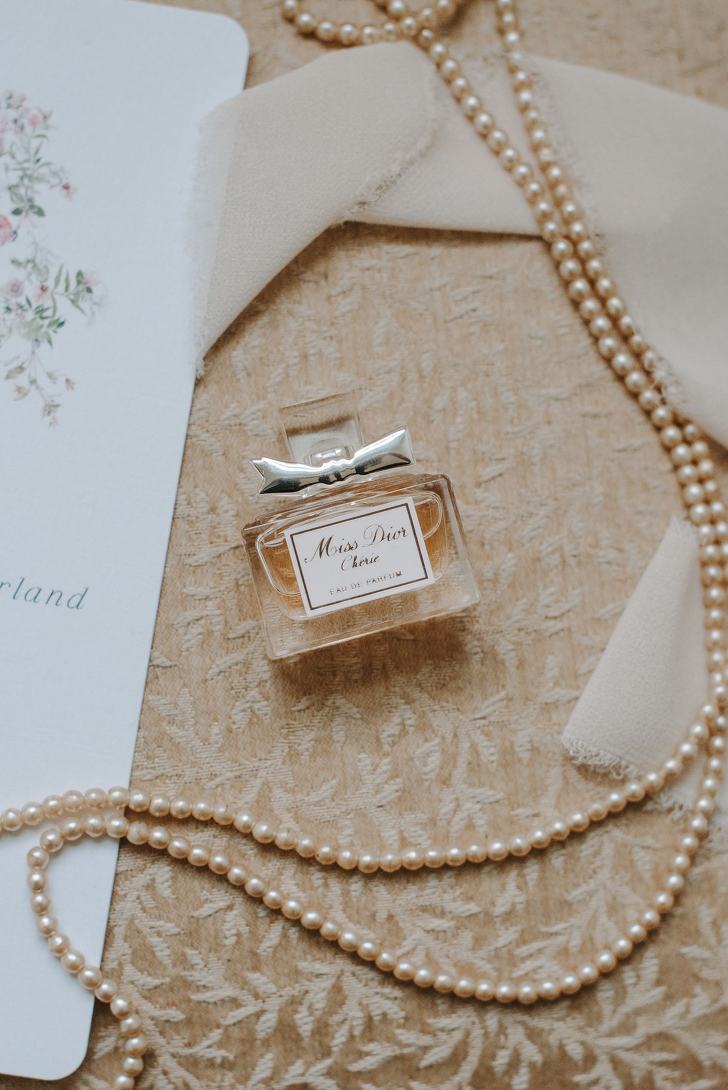 Pearl necklace and Dior perfume bottle arranged on textured beige surface with delicate paper details.