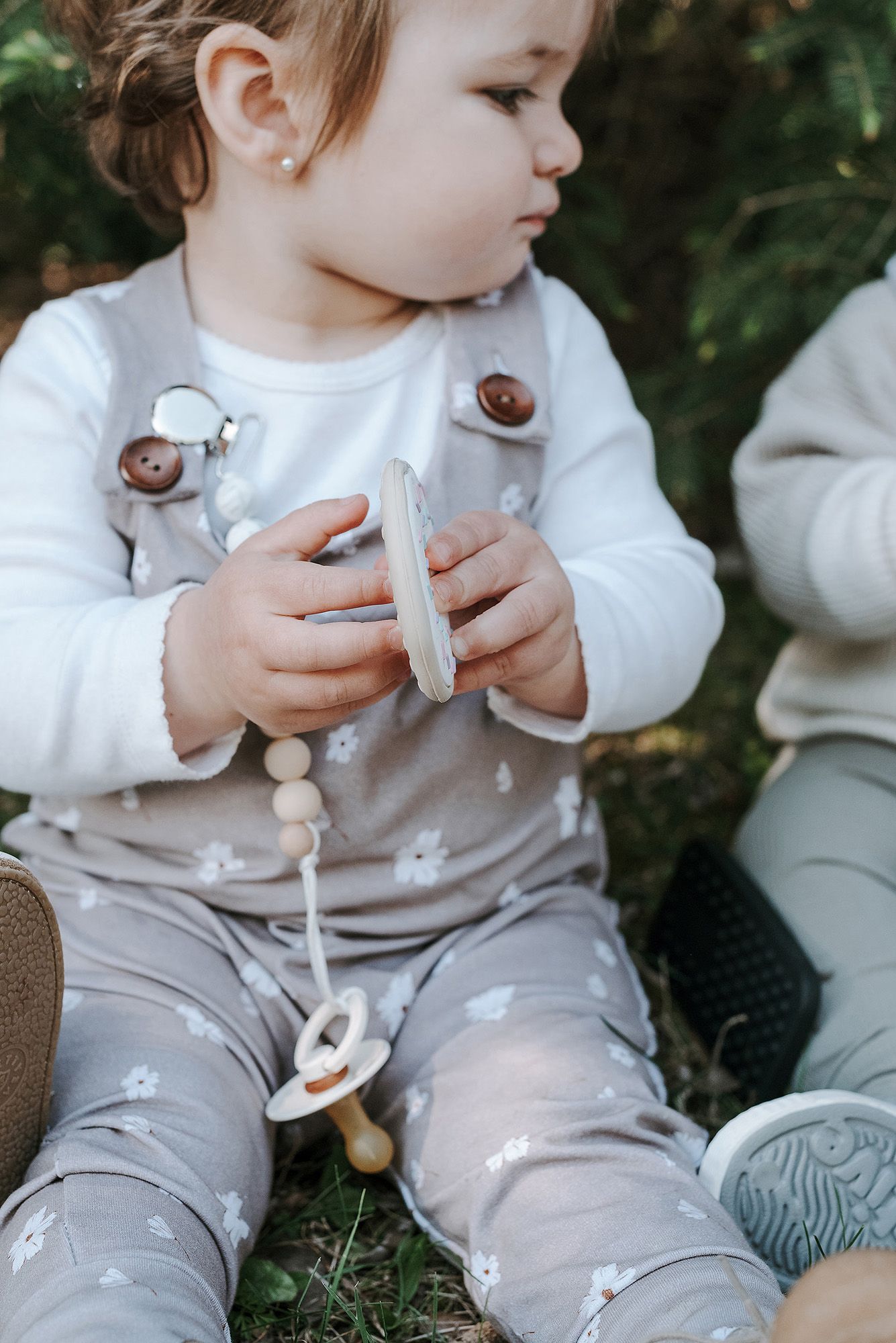 A close-up of a toddler in gray overalls holding something while sitting outdoors.
