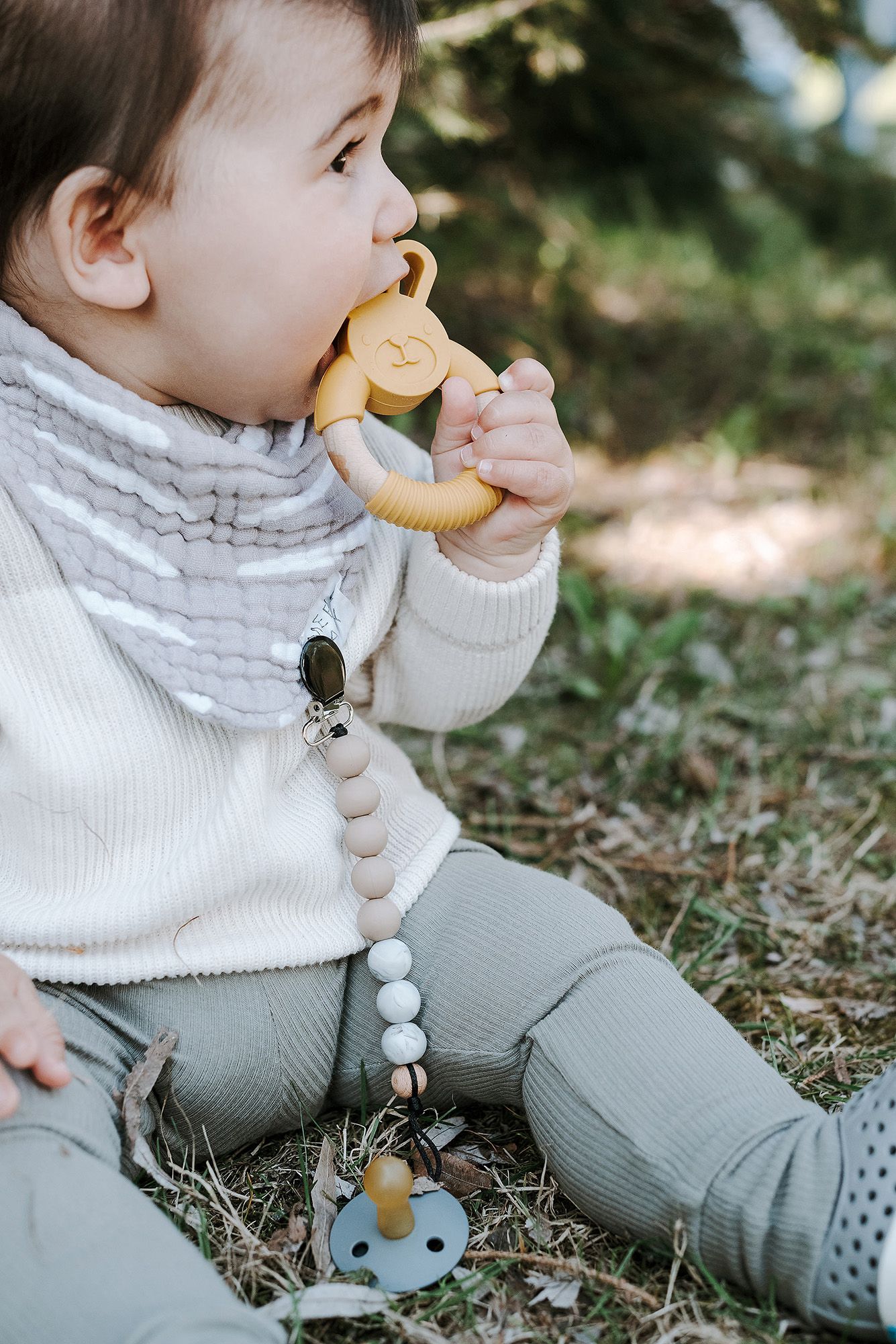 Baby in white sweater sits on grass playing with a wooden teething toy and pacifier clip.