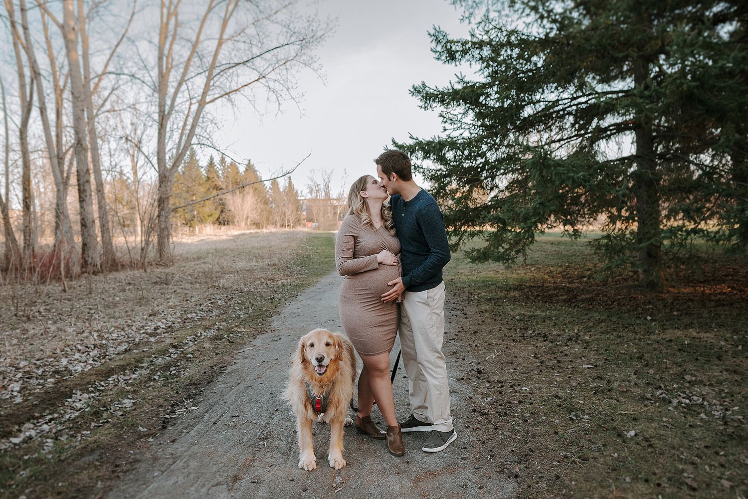 A couple walks their golden retriever through a scenic wooded path on an autumn day.