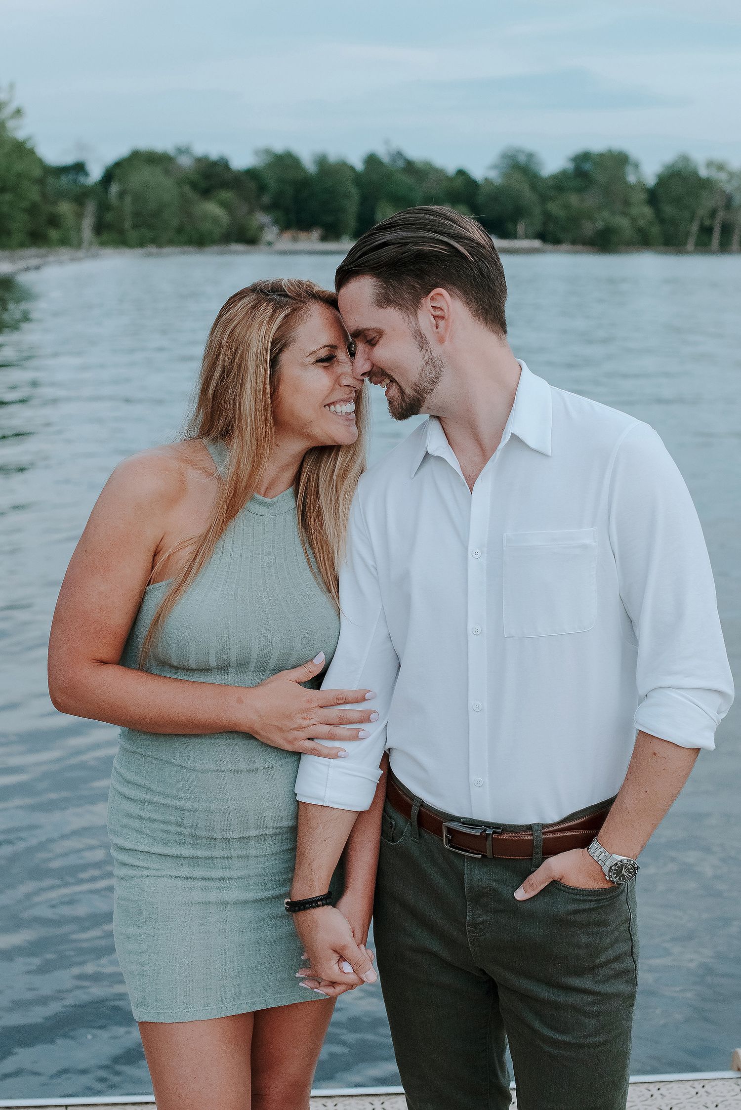 A romantic couple shares intimate moments by a lake on a summer evening, dressed in white and sage green outfits.