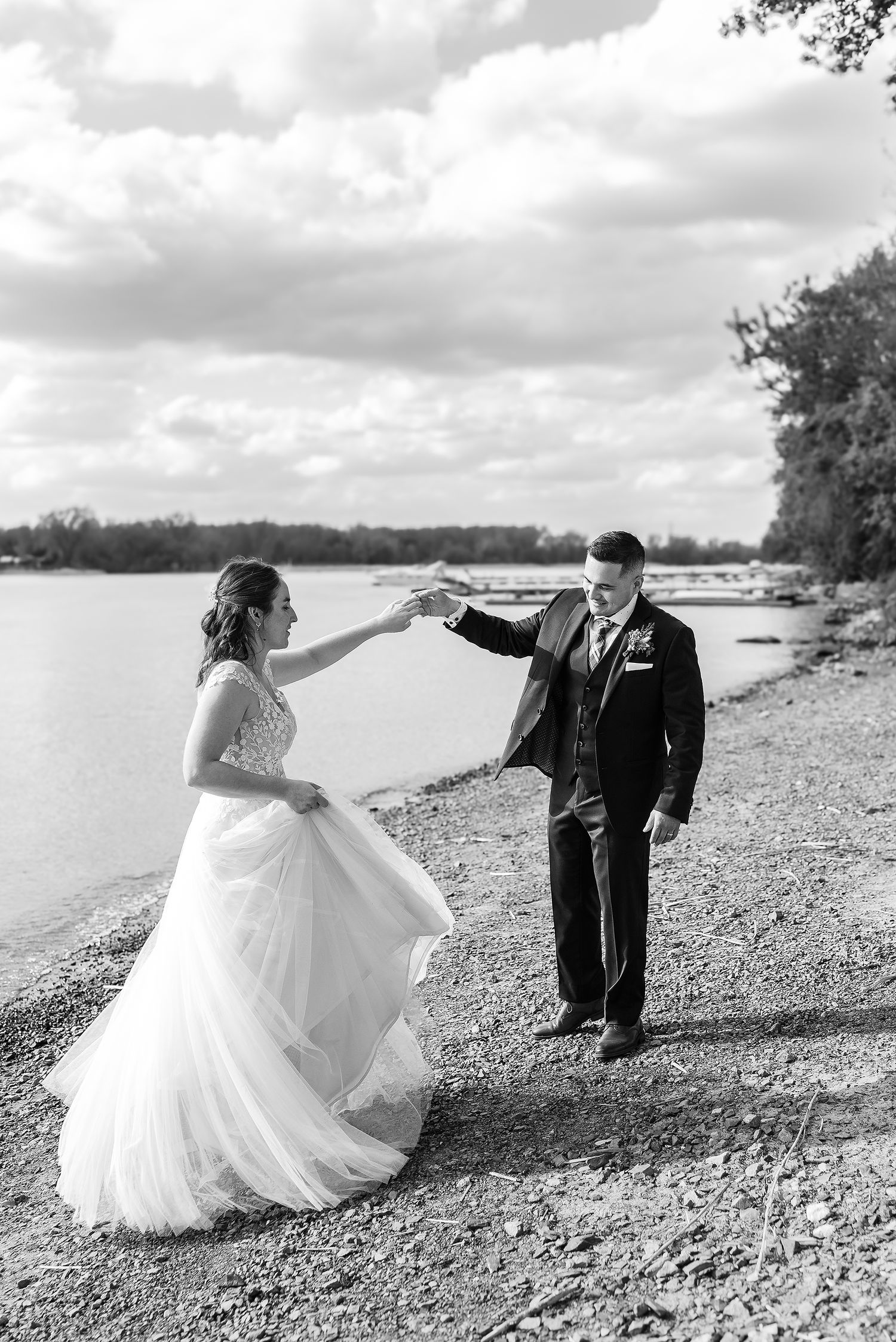 Black and white wedding couple dancing playfully on a rocky beach shore with water and trees in background.