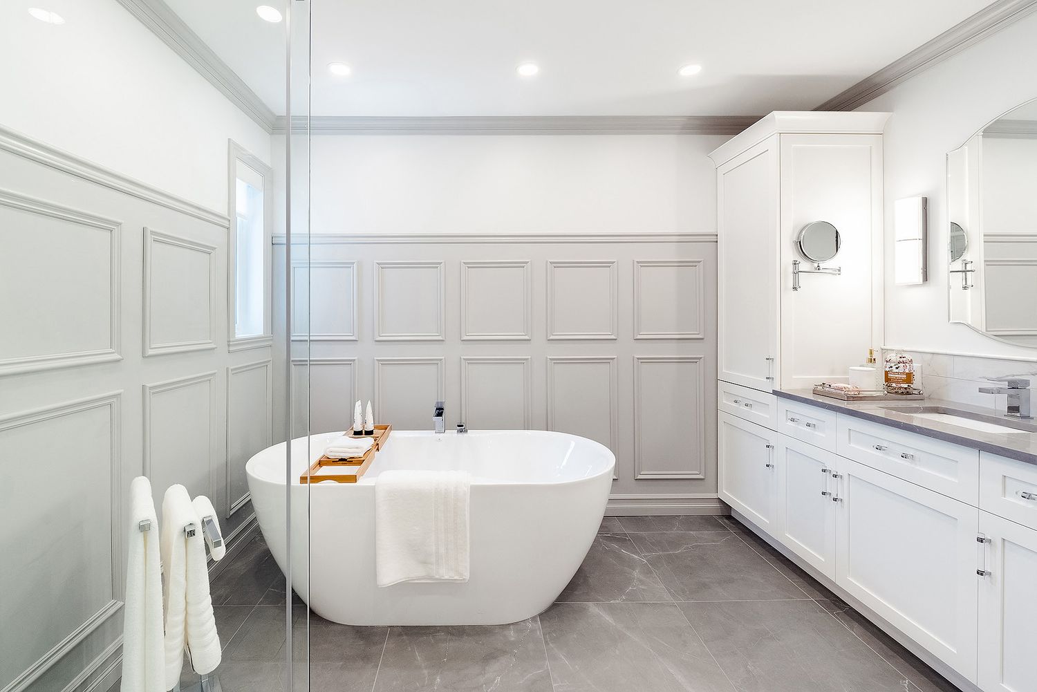 Modern white bathroom with freestanding tub, wainscoting, and marble floors features elegant traditional design elements.