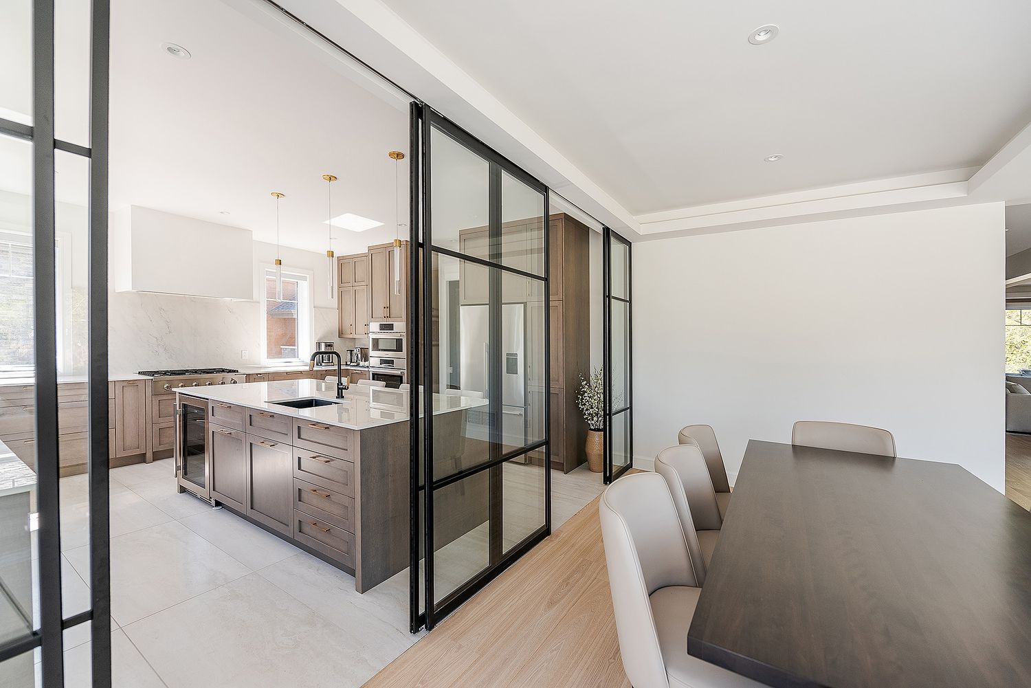 Modern kitchen with black-framed glass partition wall separating dining area featuring sleek cabinetry and wood floors.