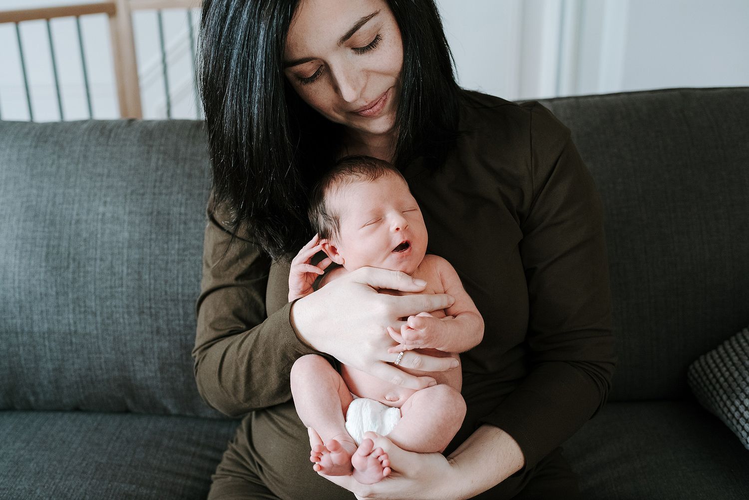Tender moments between parent and newborn on a gray couch in natural window light.