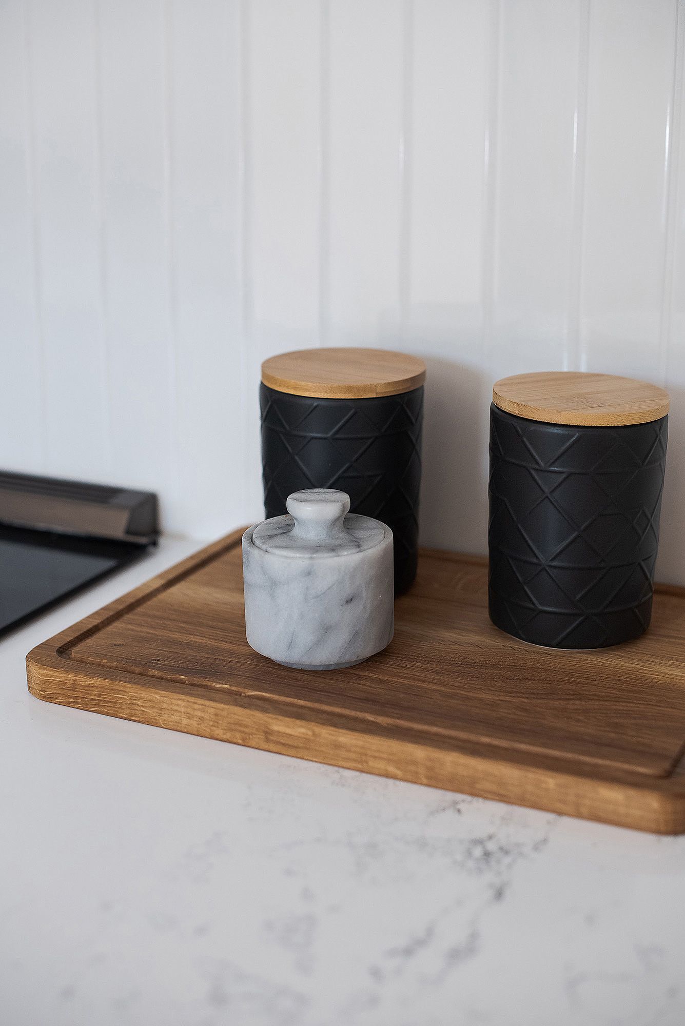 Black ceramic storage containers with wooden lids arranged on a rustic wooden serving board.