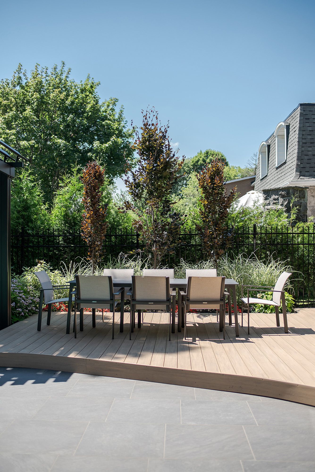 Modern outdoor patio with composite decking, dining furniture and lush landscaping against a bright blue sky.