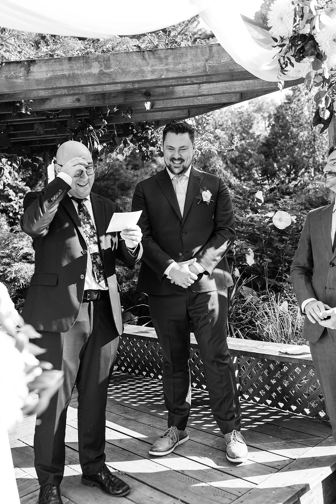 A group of groomsmen in formal black suits sharing drinks and laughter at an outdoor wedding reception.