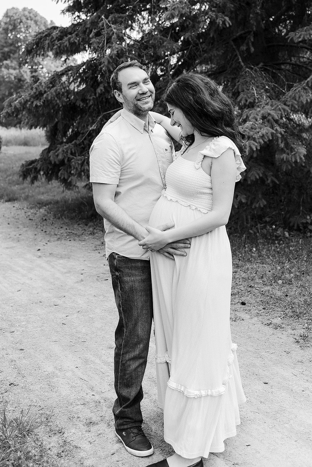 A couple in casual white outfits embracing and laughing together on a dirt path surrounded by greenery.
