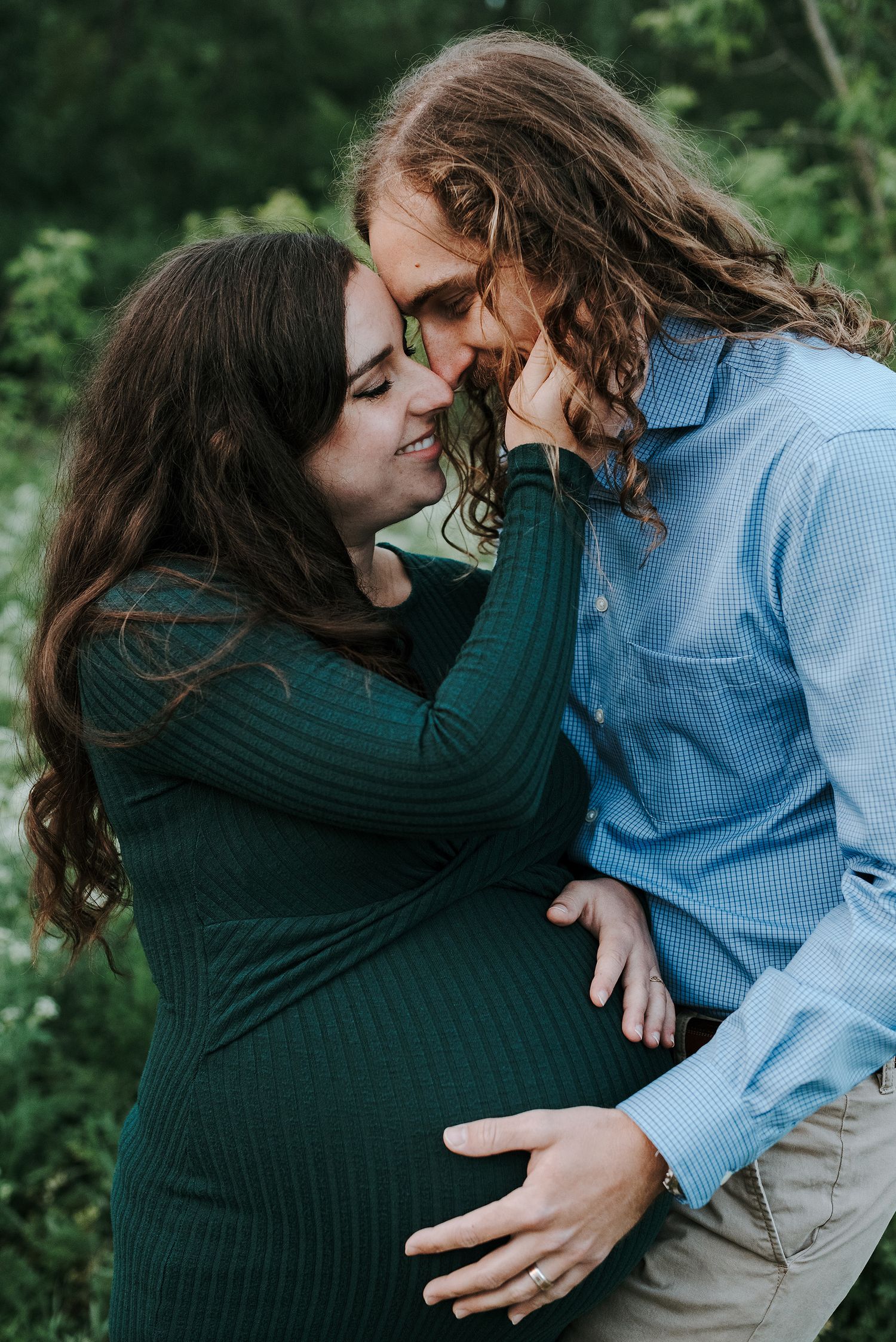 Romantic couple embracing outdoors in casual attire against lush green foliage.