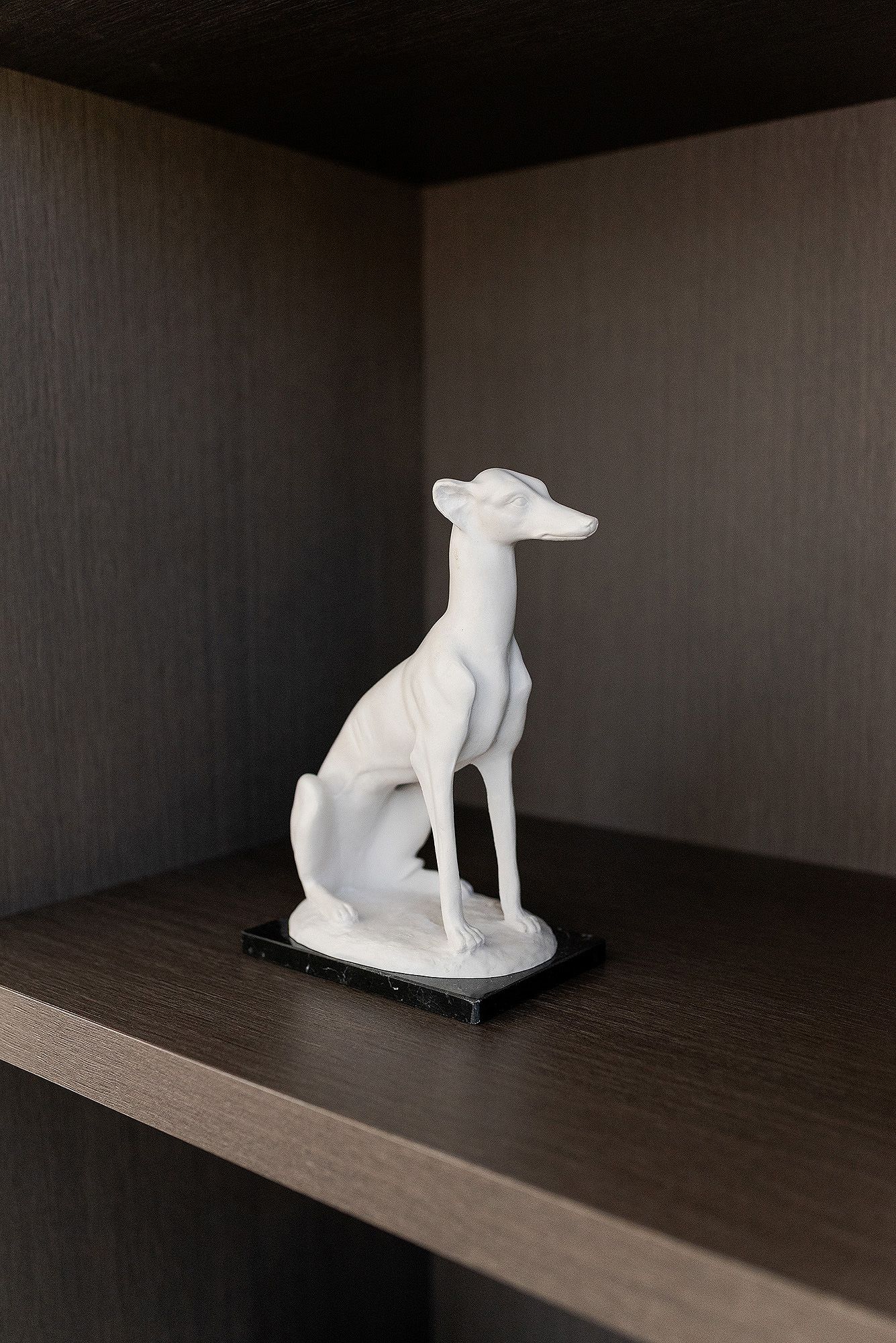 A white ceramic dog sculpture sits on a dark wooden shelf in dramatic lighting against a black background.