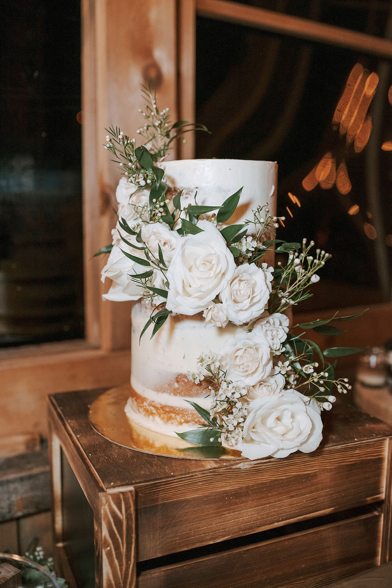 White wedding cake with fresh roses and greenery displayed on rustic wooden stand in warm lighting.
