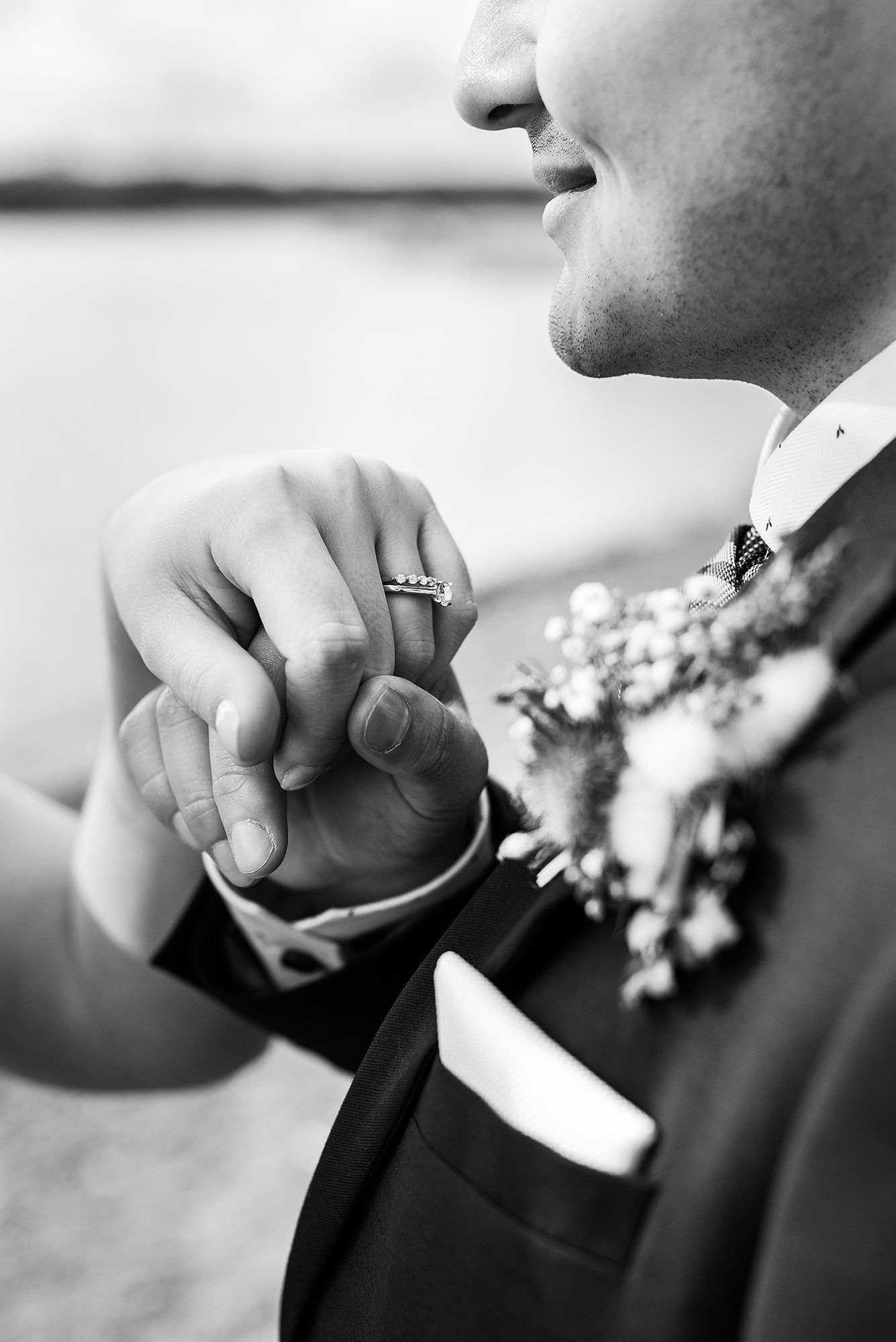 Close-up black and white photo of hands holding a wedding ring with a boutonniere visible in the frame.