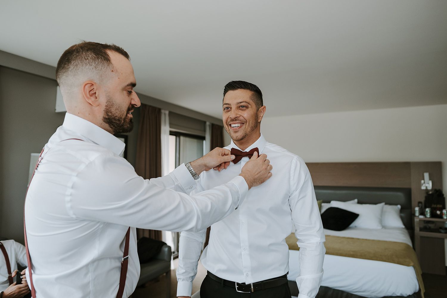 Two people in white dress shirts help each other get ready in a hotel room before a formal event.