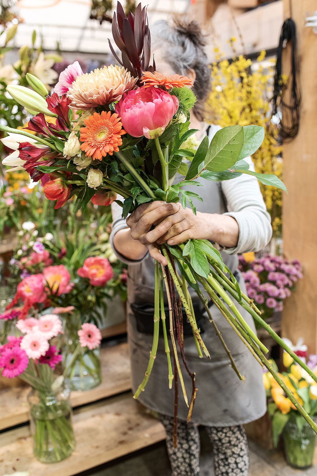 A florist arranges a vibrant bouquet with pink peonies, orange gerbera daisies and greenery in a flower shop.