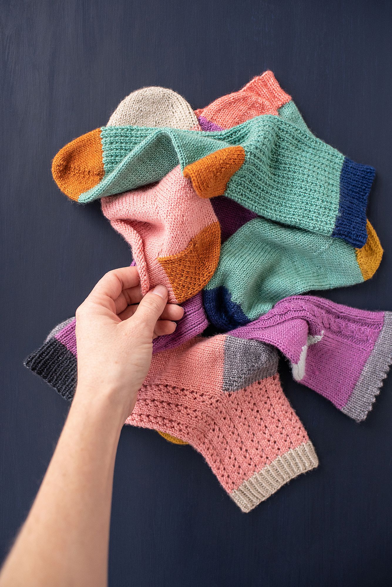 Colorful knitted socks in progress showing various shades of pink, mint green, orange and purple laid out on dark surface.