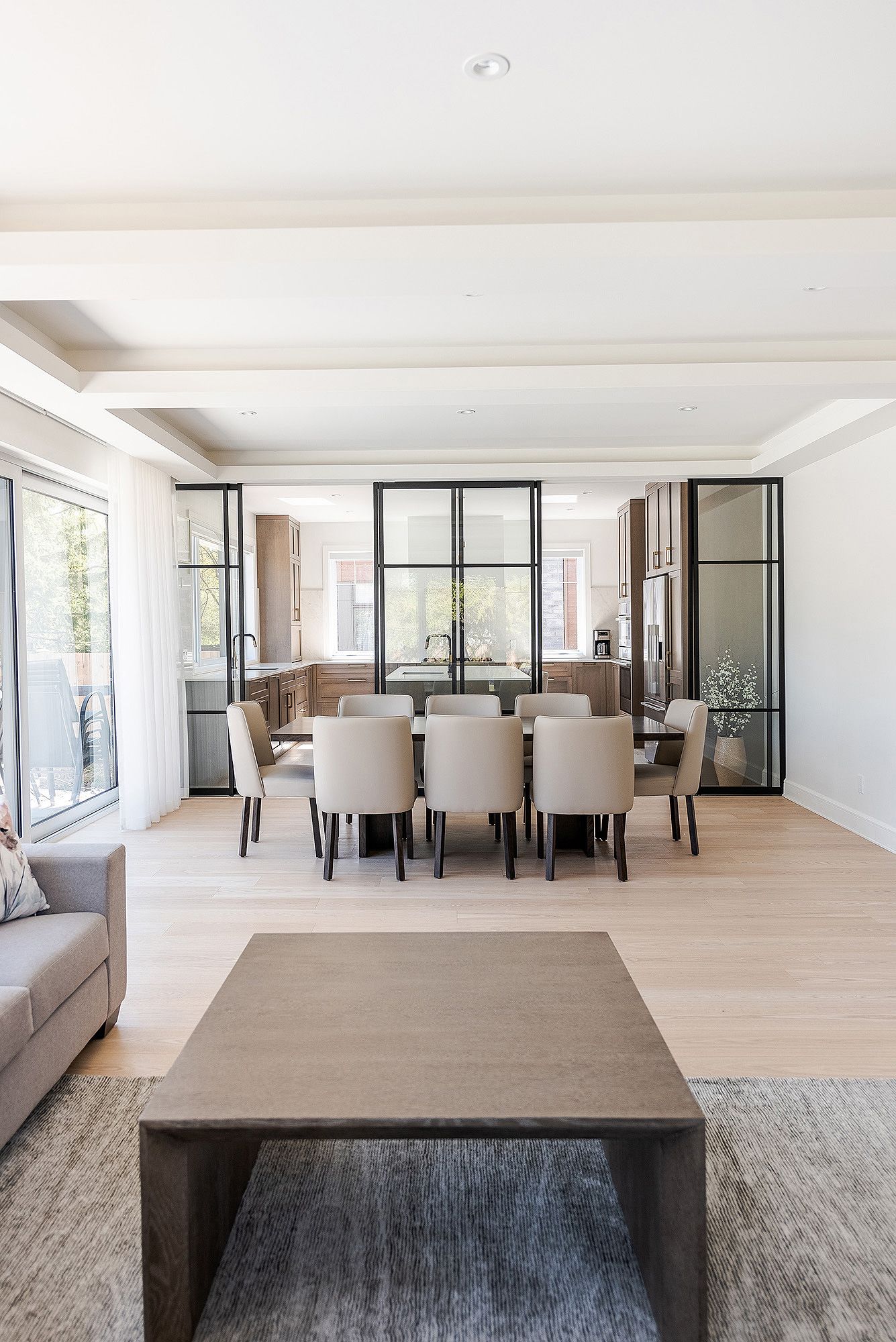 Modern open concept living and dining room with light wood floors, white walls and black framed glass partitions.