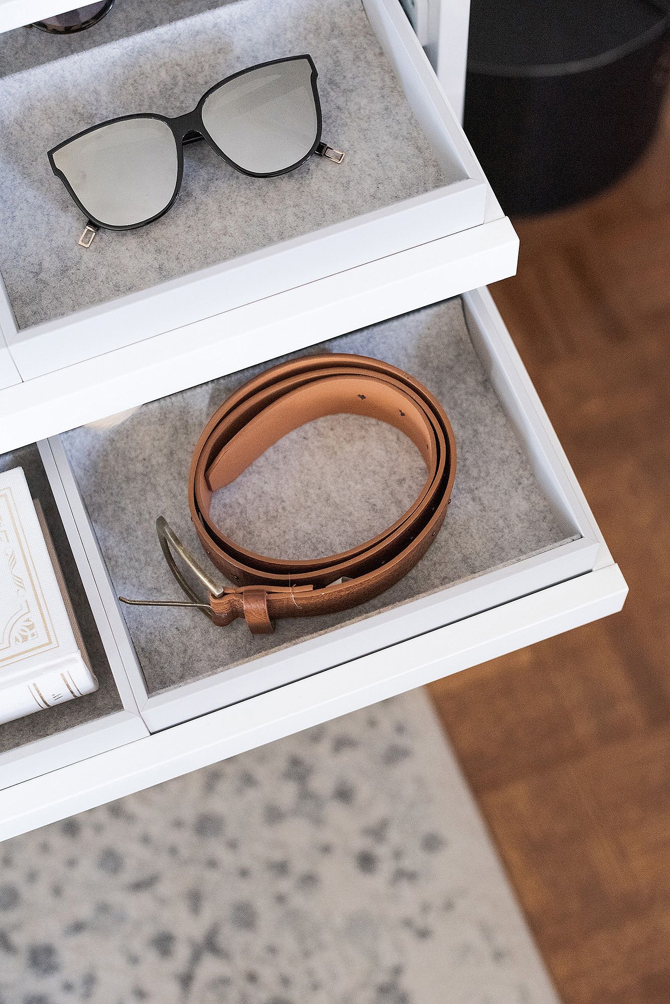 Sunglasses and brown leather belt arranged neatly in white storage drawers viewed from above.