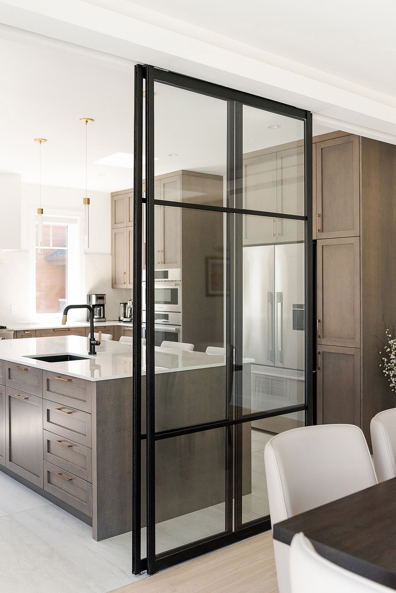 Contemporary kitchen with black-framed glass divider, taupe cabinets, and pendant lighting.