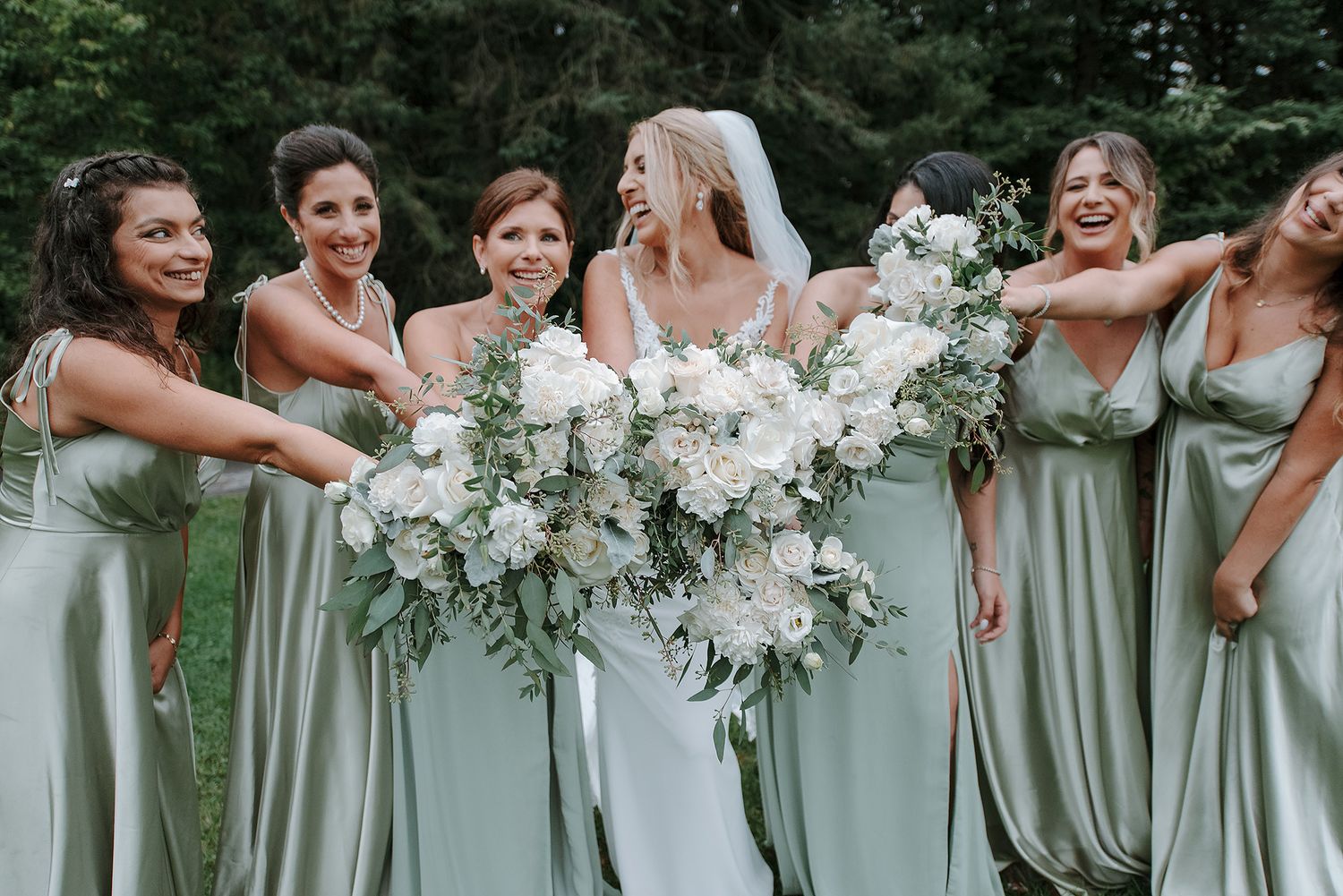 A group of bridesmaids in sage green dresses hold white floral bouquets while posing together outdoors.