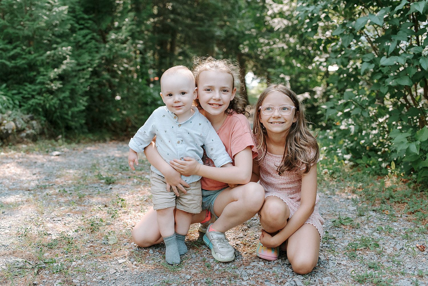 A family in casual summer outfits poses together outdoors in a wooded setting during a photo session.