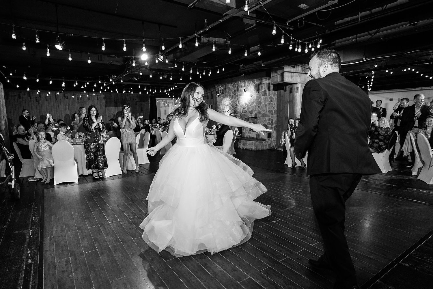 Newlyweds share their first dance on a rustic wooden dance floor under twinkling string lights at an evening reception.