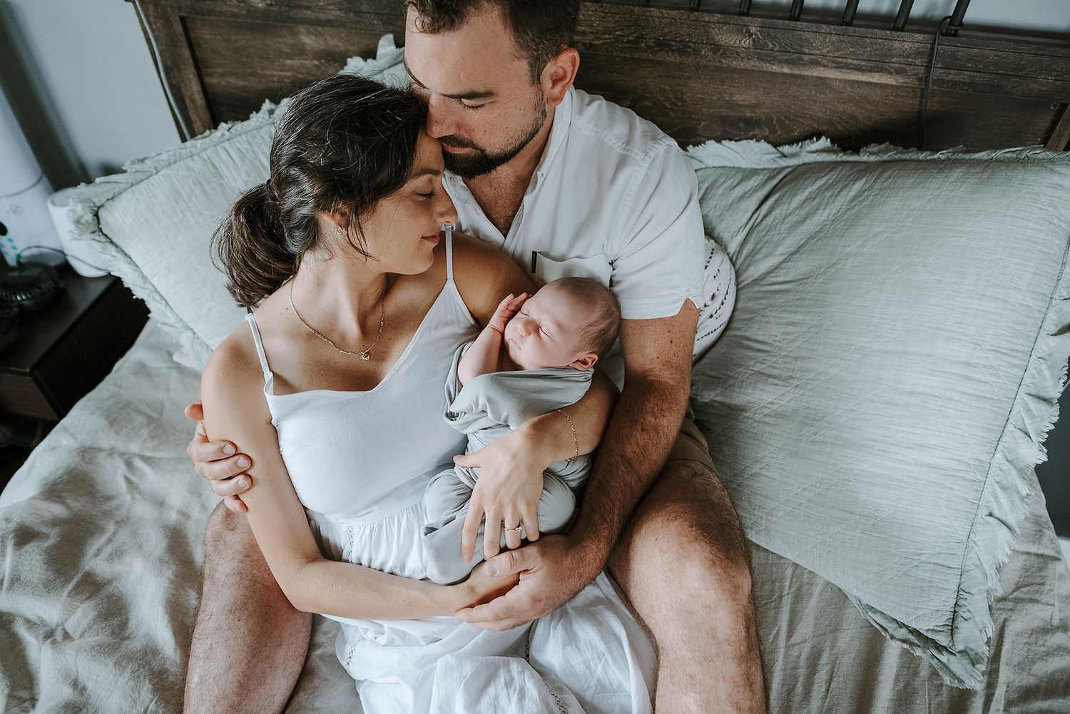 An intimate bedroom scene of parents cuddling with their newborn baby on white bedding.