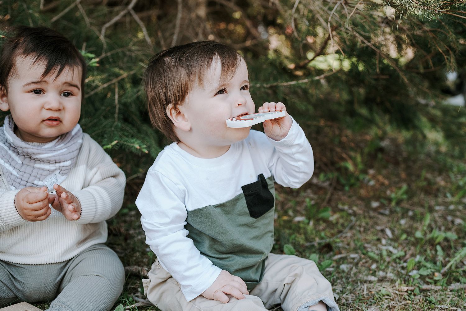 Two toddlers in casual wear sit together outdoors while enjoying snacks.