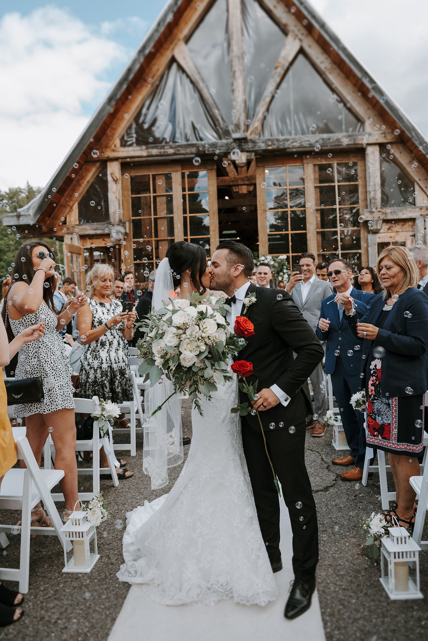 Wedding guests celebrate at a rustic timber-framed venue with white chairs and floral decorations.