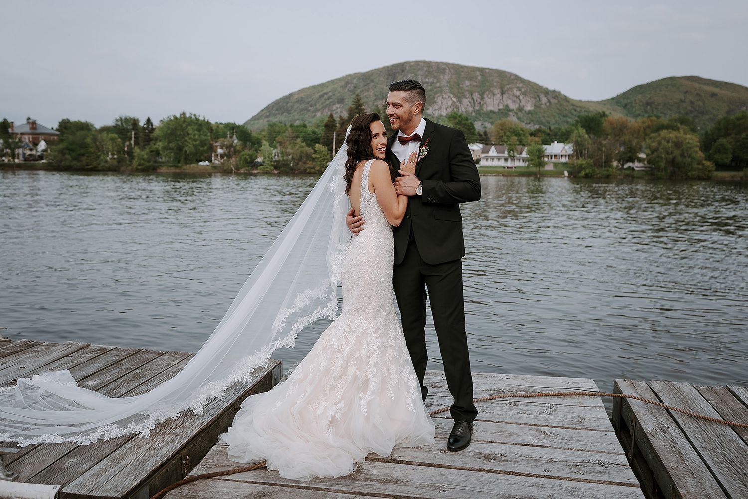 A couple in wedding attire walks together on a wooden dock by a serene lake with mountains in the background.