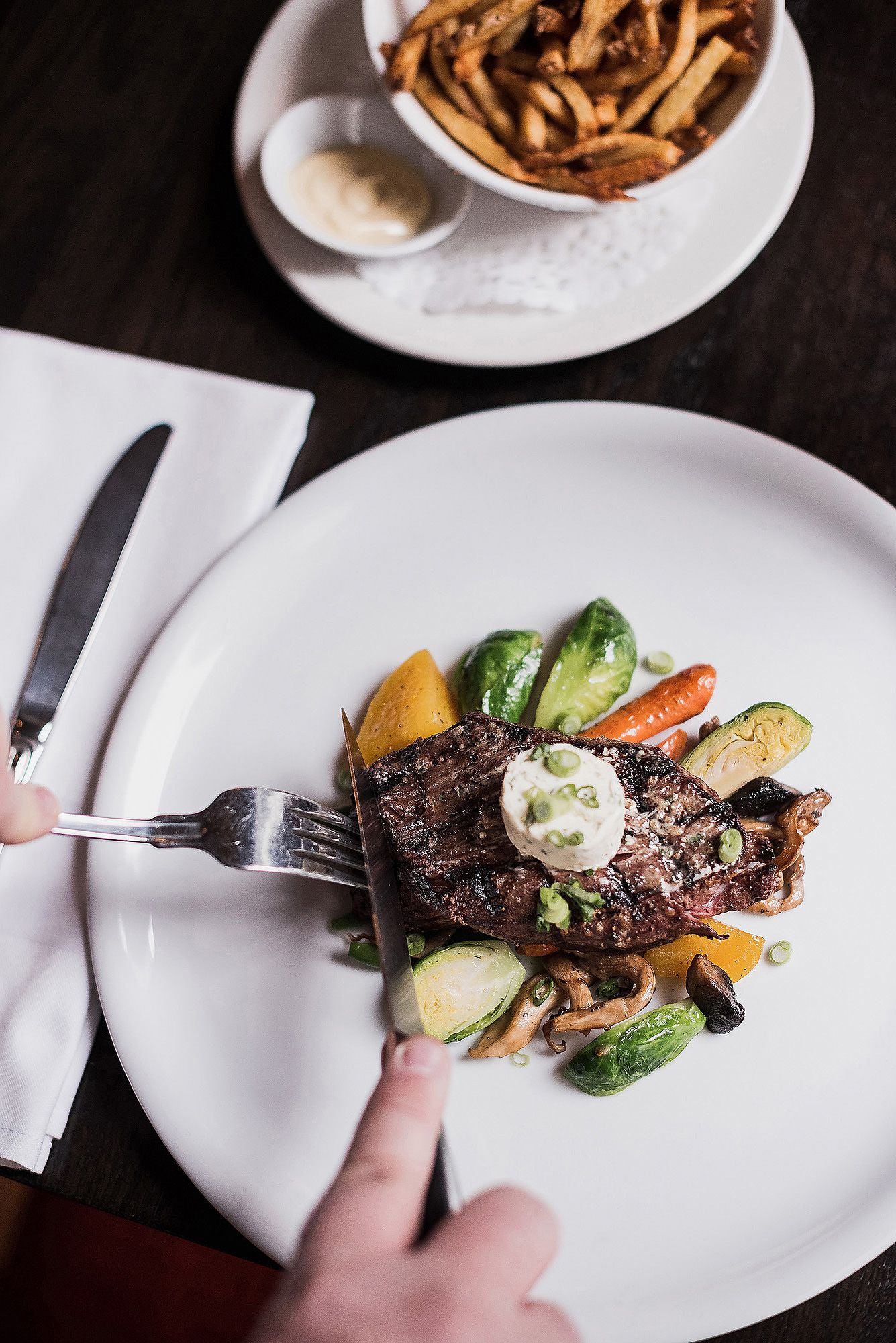 Grilled steak entree served with roasted vegetables and compound butter on white plates in an upscale restaurant setting.