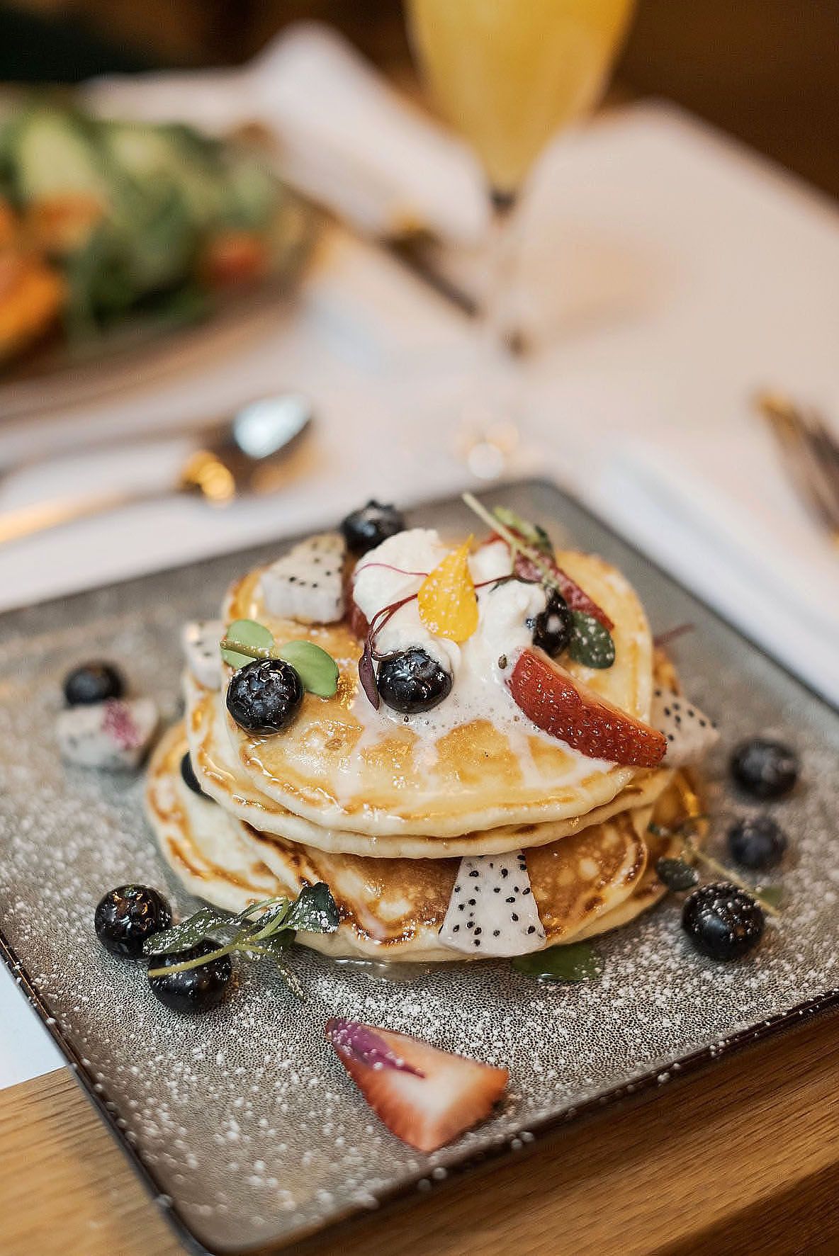 Stack of pancakes topped with fresh berries, powdered sugar, and fruit on a grey slate plate.
