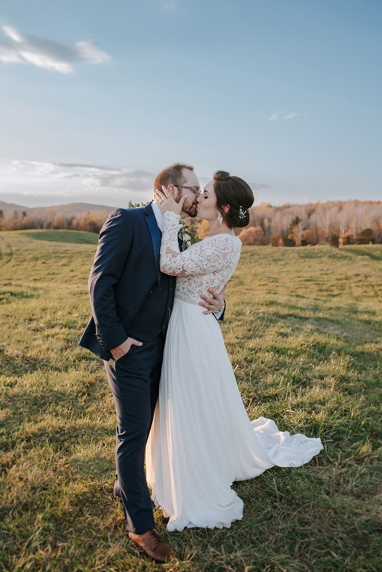 A romantic wedding couple embraces in a sunlit meadow with rolling hills and autumn trees in the background.