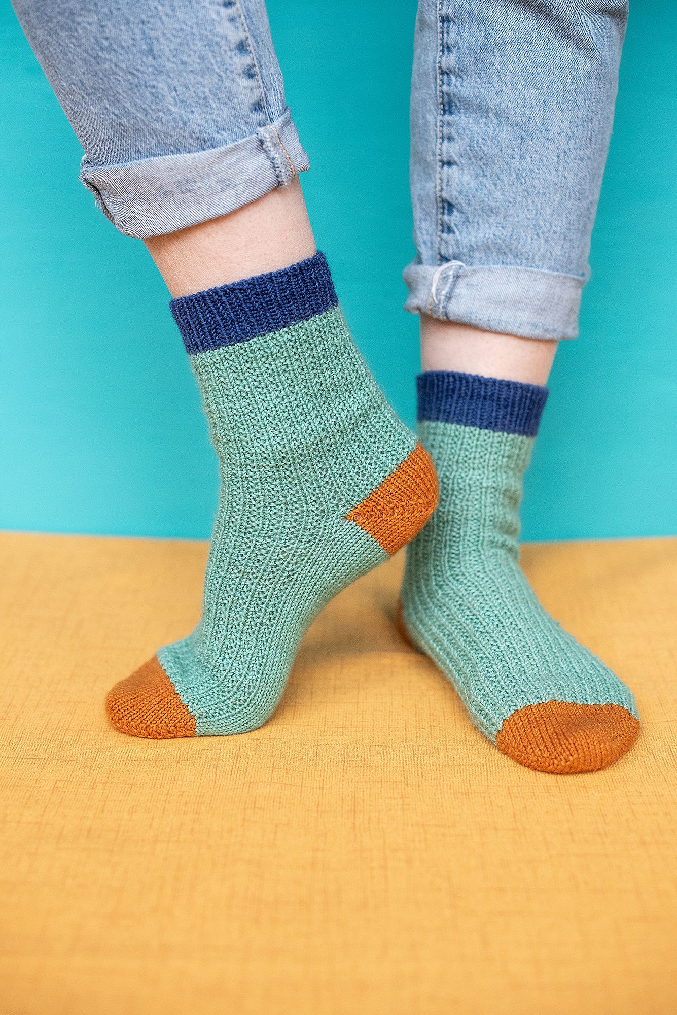 Mint green knitted socks with orange toes and blue ankles against turquoise and yellow background.