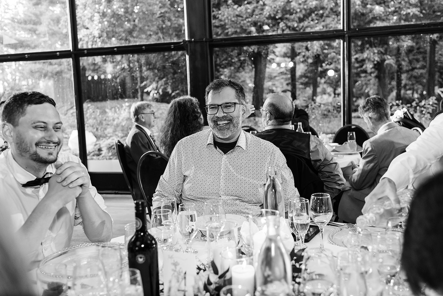 Black and white photo of guests enjoying drinks and conversation at a dinner reception with wine glasses on the table.