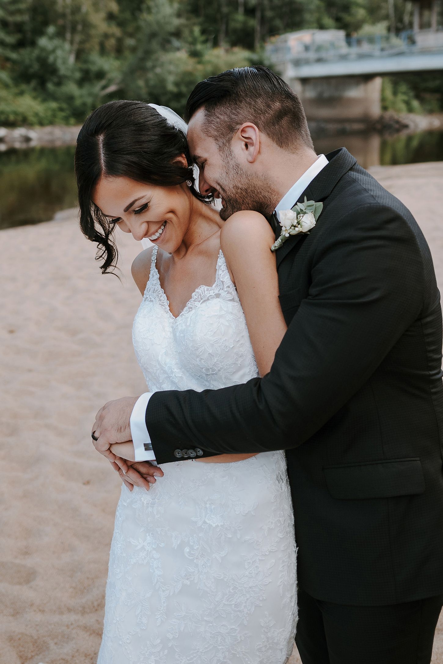 A tender wedding moment captured outdoors as a couple embraces and shares a joyful laugh by a lakeside.