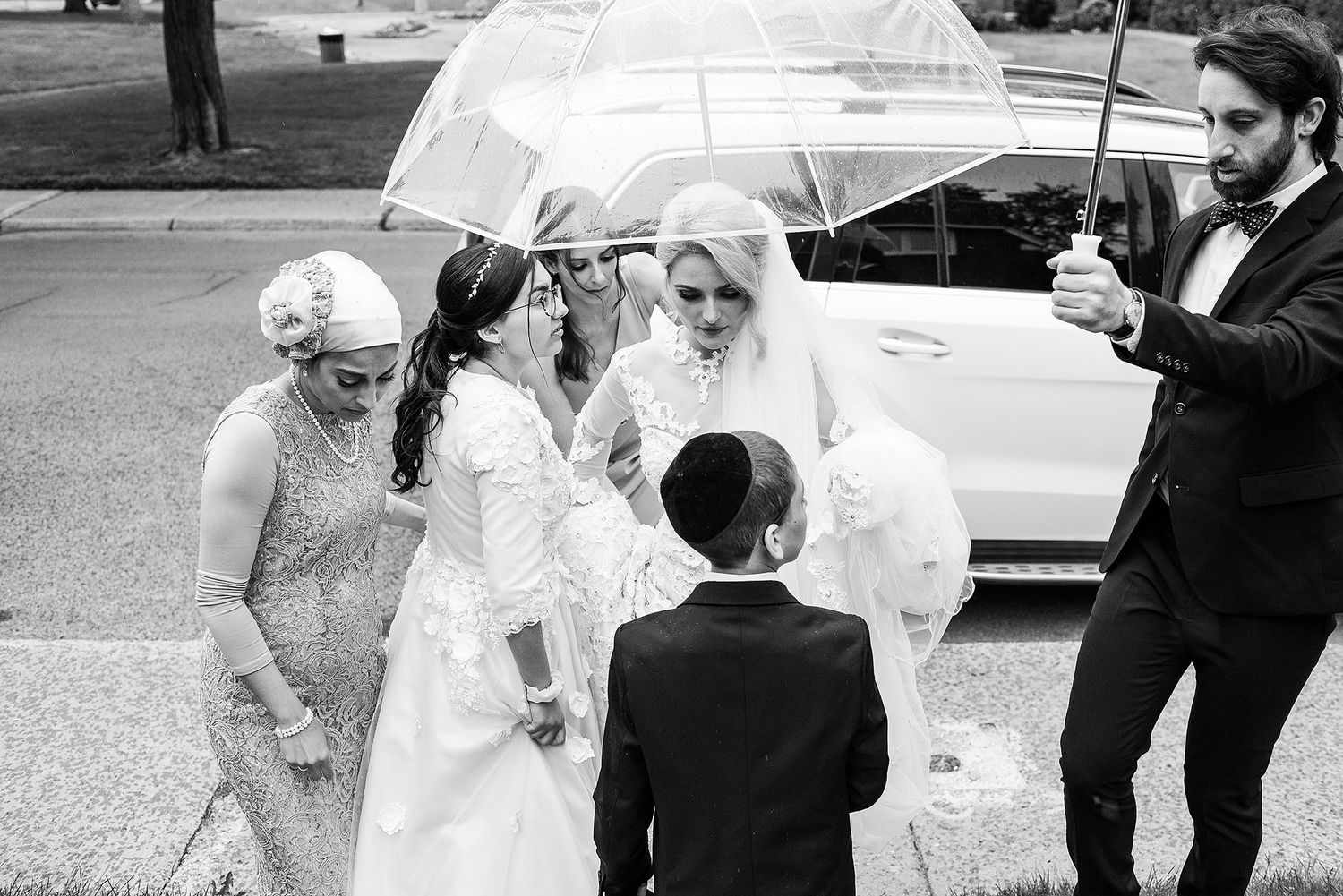 Wedding party gathers under white umbrellas on a rainy day outside near parked cars.