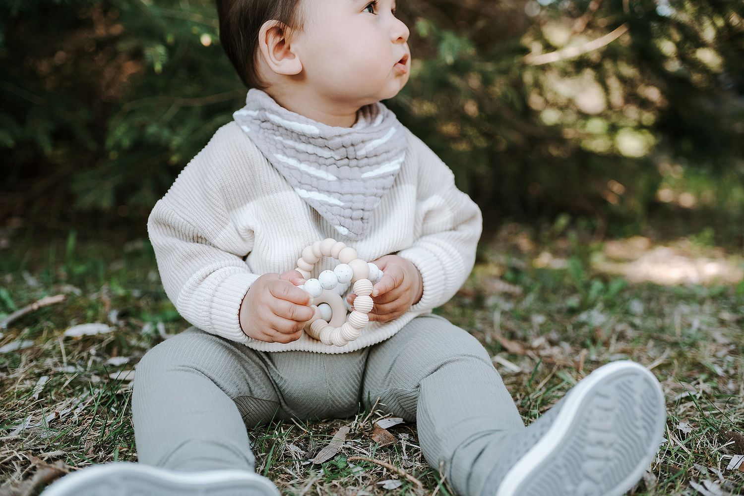 Baby in white sweater and gray pants sitting thoughtfully on ground outdoors.
