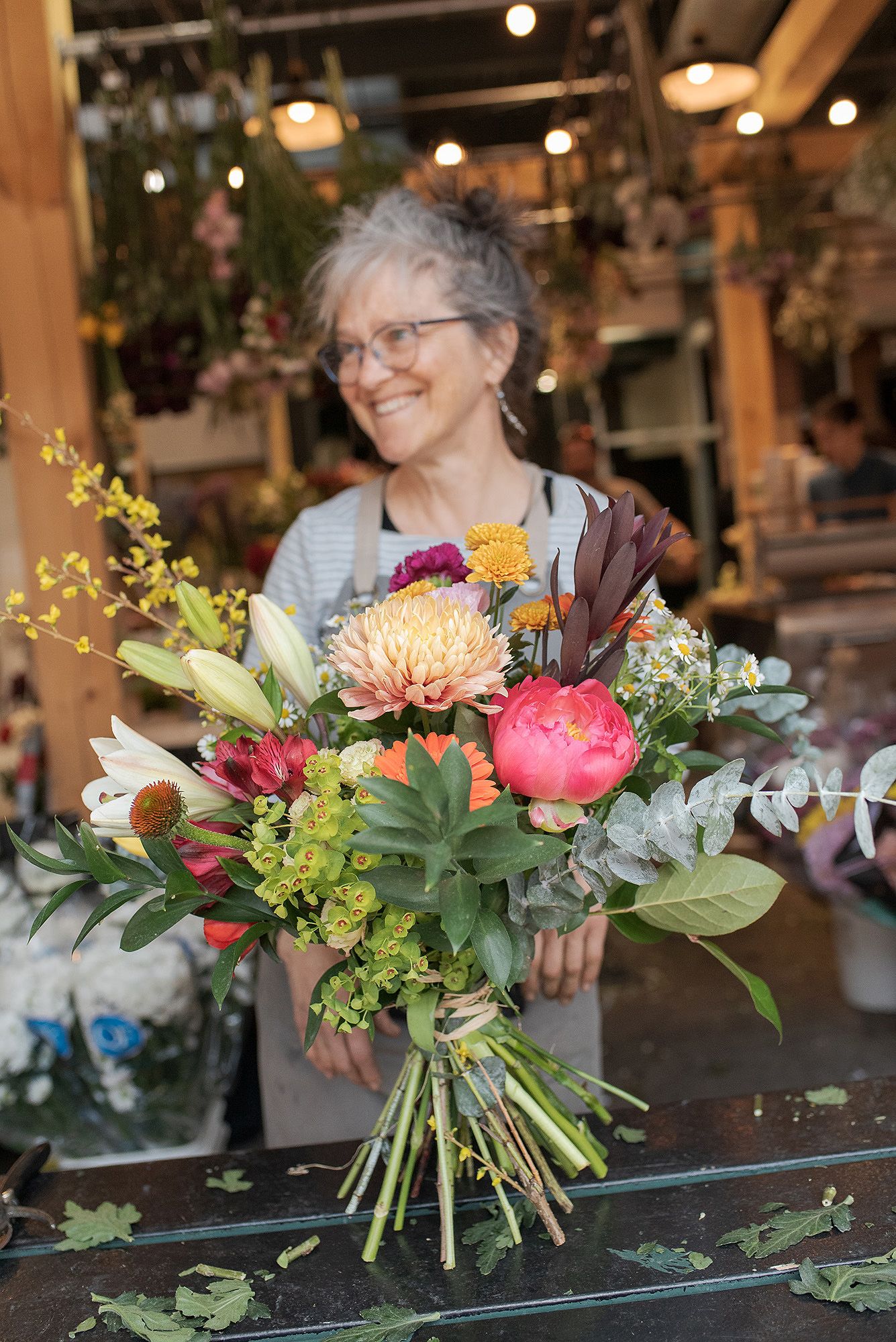 A vibrant floral bouquet with pink peonies, yellow chrysanthemums and greenery sits on a glass surface in a flower shop.