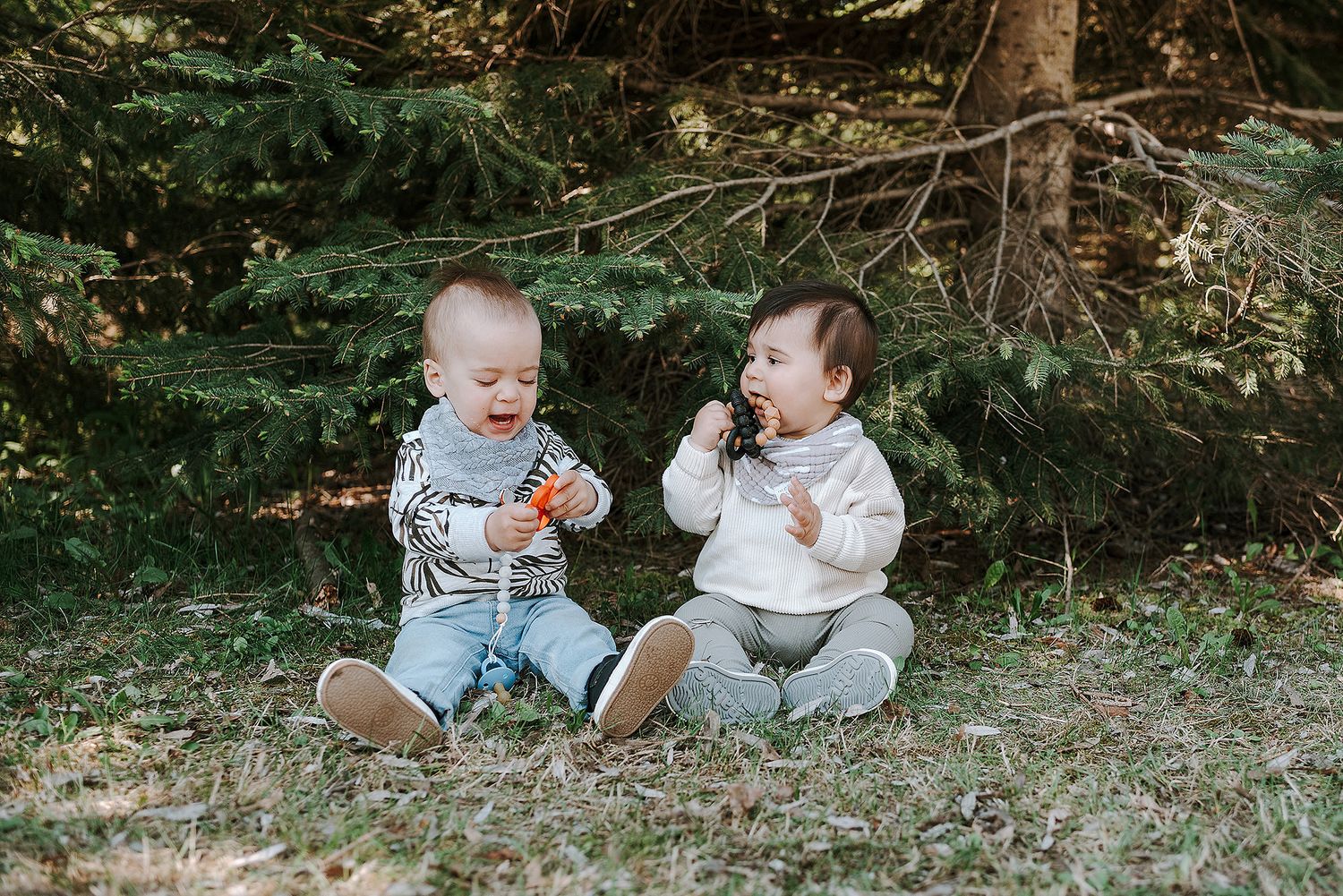 Two babies sitting on grass playing together in a wooded outdoor setting.