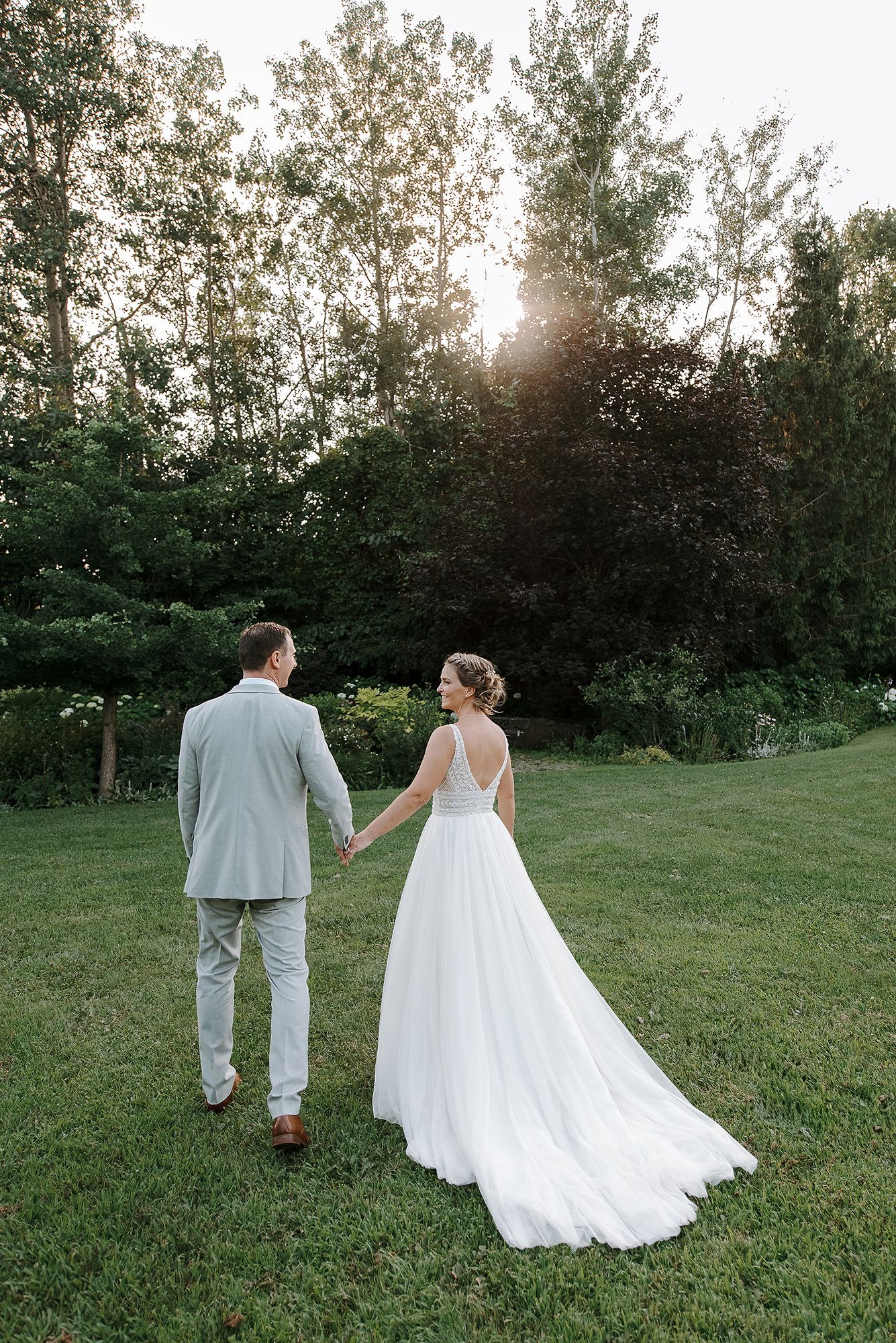 A couple in wedding attire walks hand in hand through a grassy field at sunset with trees in the background.