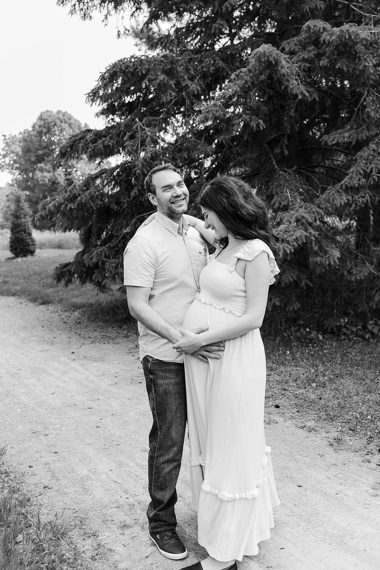 A couple in casual attire embraces on a dirt path surrounded by lush green trees in a romantic outdoor setting.