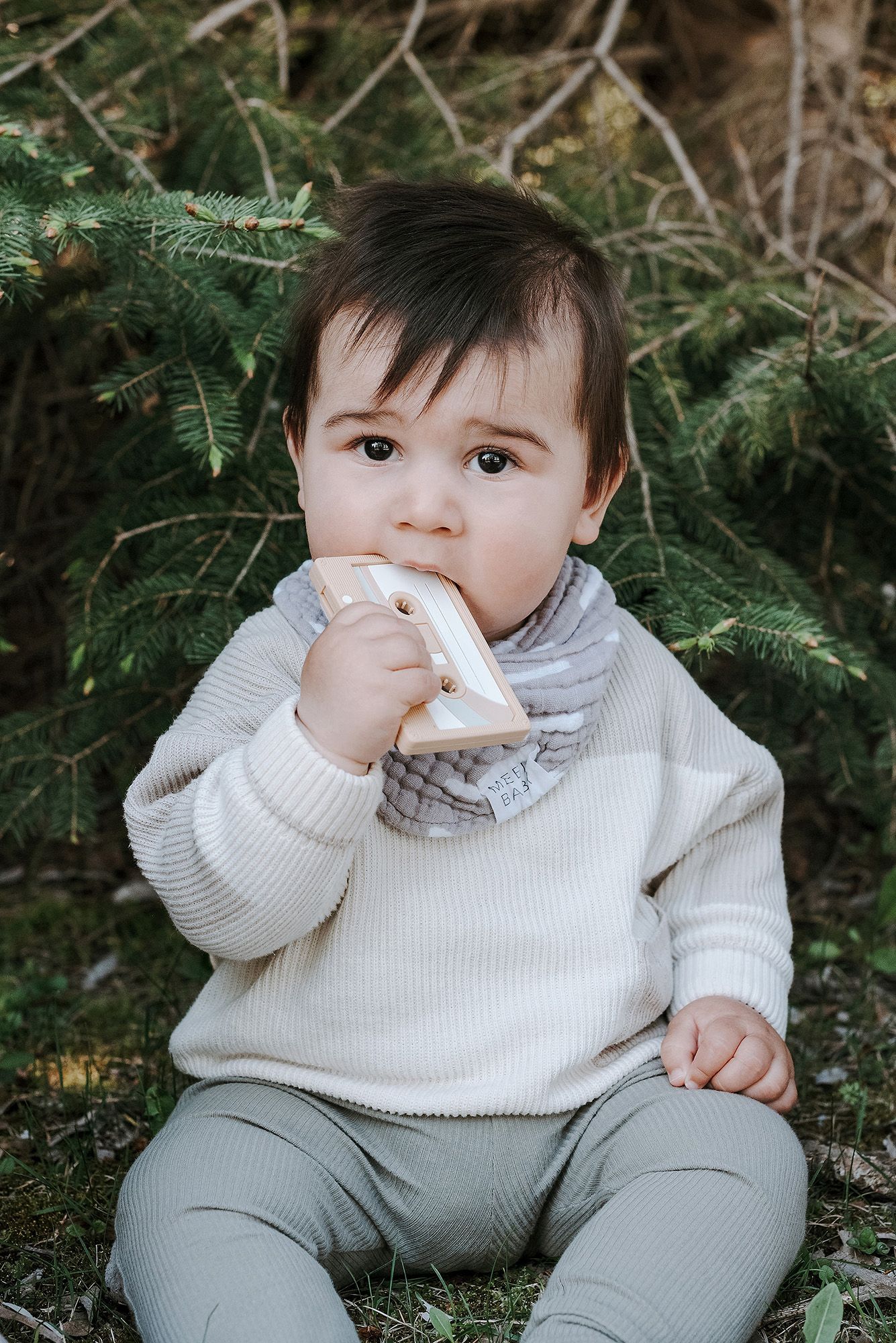 A small child in a white sweater sits against evergreen branches while holding something to their mouth.