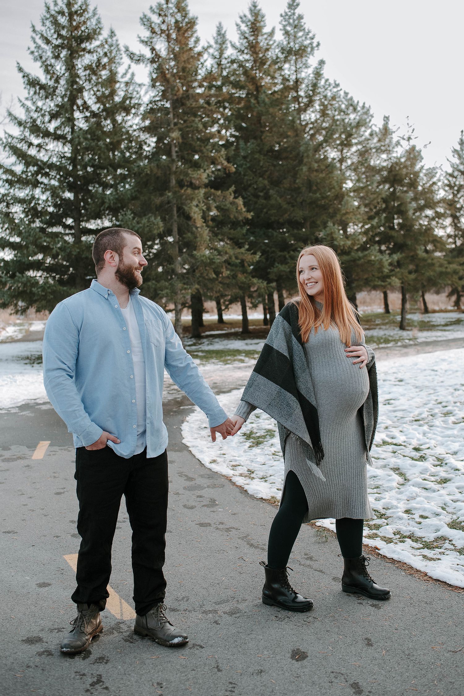 A couple stands together in a snowy park with evergreen trees, the woman wearing a gray wrap.