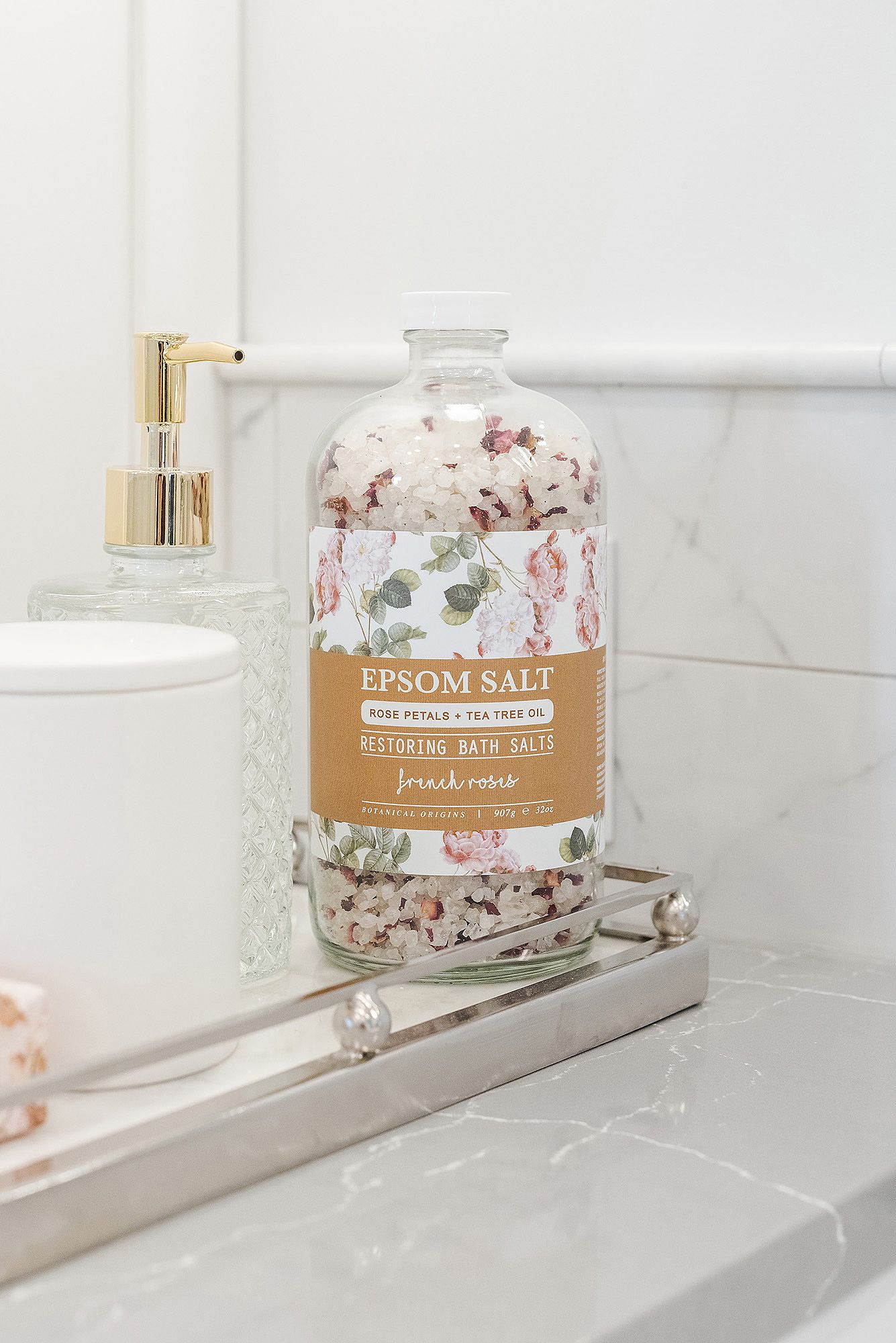 Glass jar with floral pattern and gold lid containing epsom salt sits on white marble bathroom shelf.