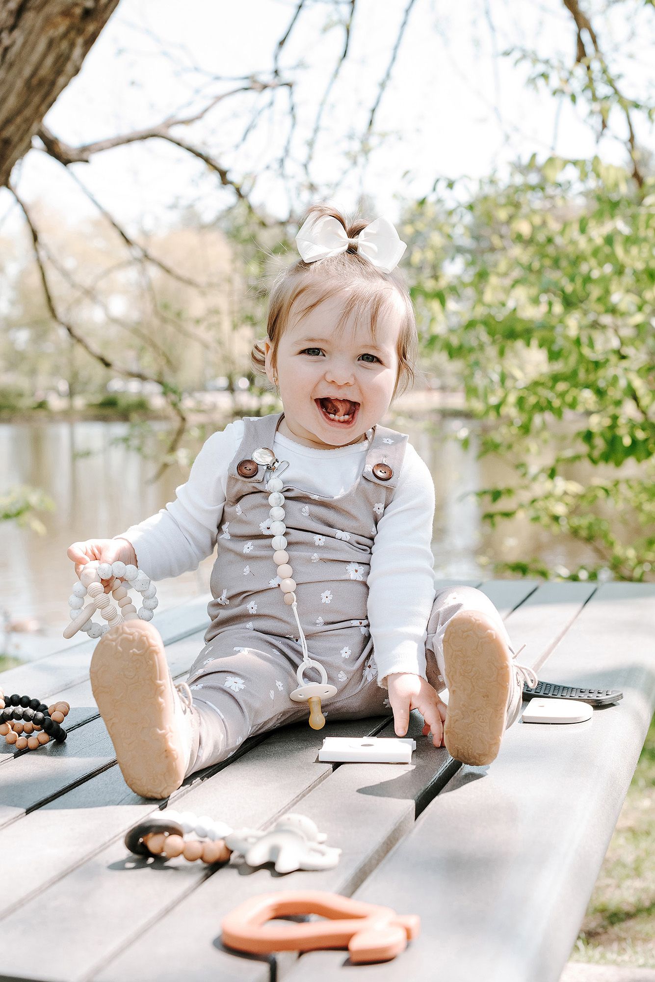 Adorable baby in gray overalls laughing while sitting on a wooden bench outdoors.