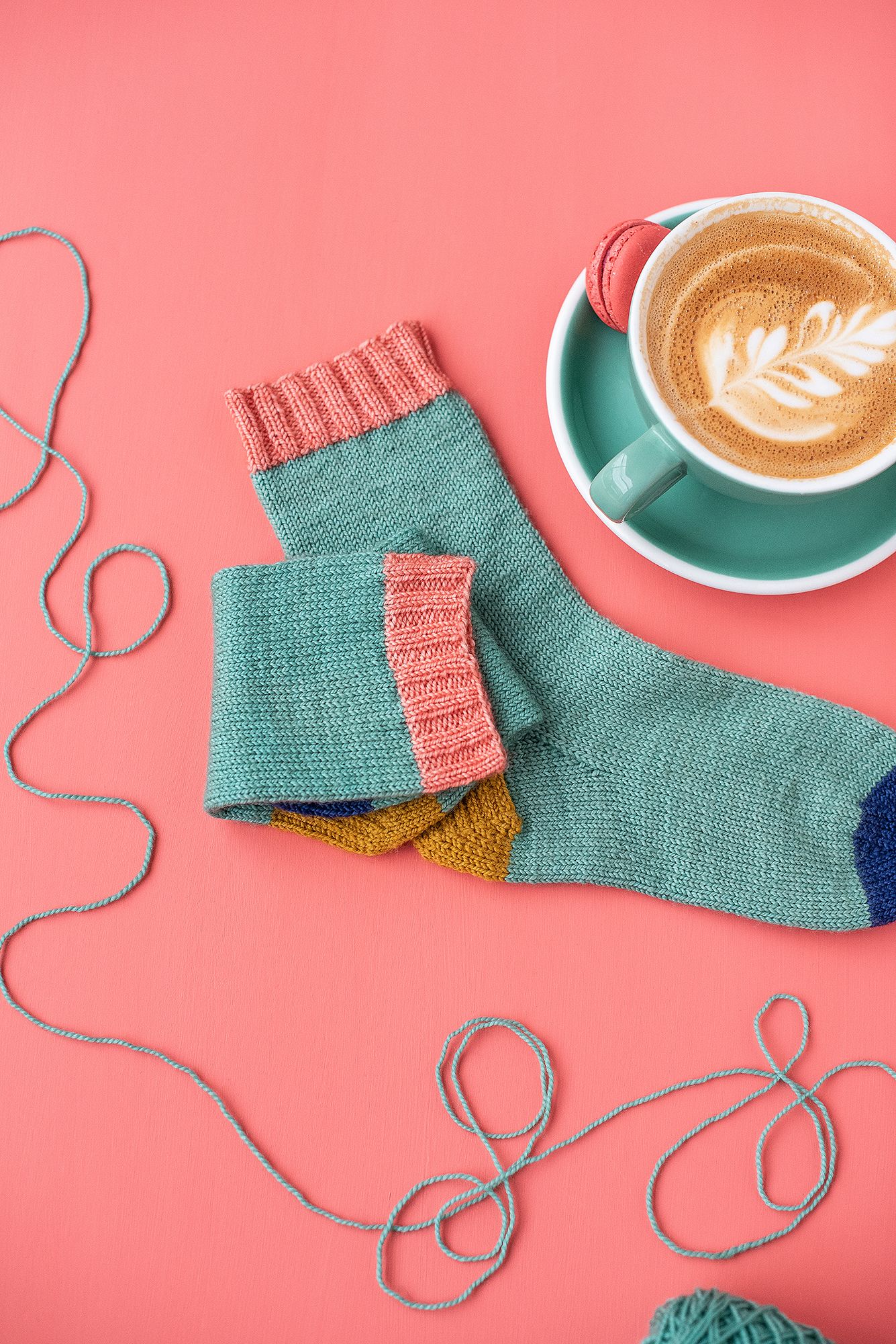 Knitted socks and coffee with latte art on coral background with knitting supplies.