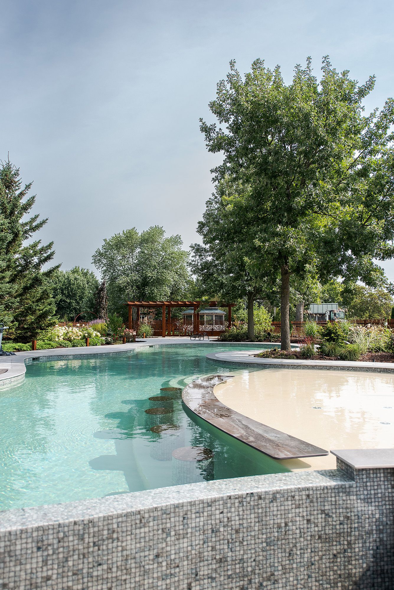 Luxurious curved swimming pool with mosaic tile detail and landscaped garden features a modern pergola in the background.