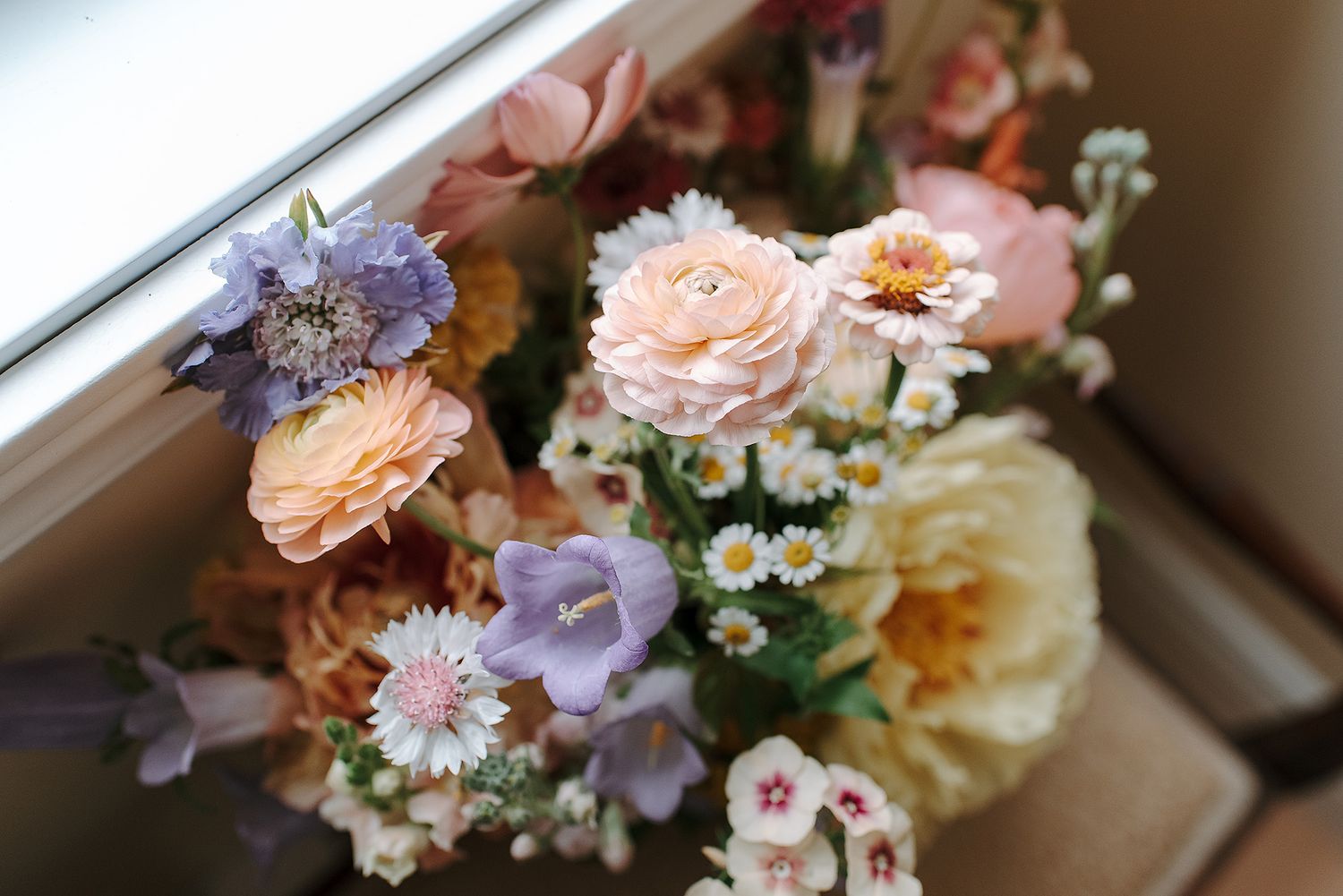 A lush bouquet of pastel flowers including ranunculus, daisies, and bluebells arranged near a sunlit window.